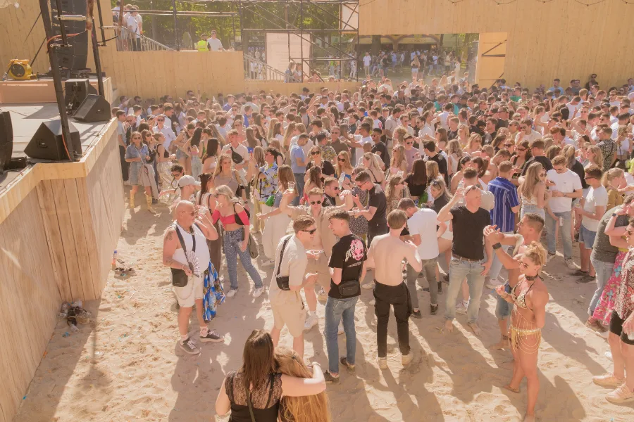 A crowd dancing outdoors in daylight on a sandy lot beside a stage